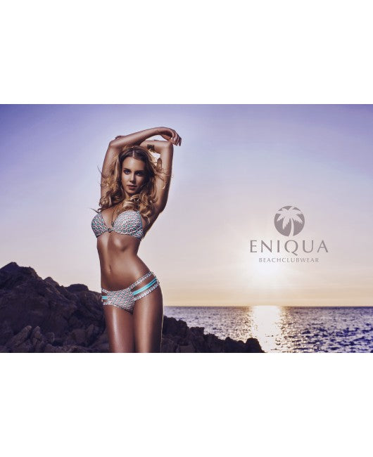 A woman in a patterned push-up bikini poses at sunset on a rocky shore, next to her the Eniqua logo, which underlines the charm of Italian Eniqua swimwear.